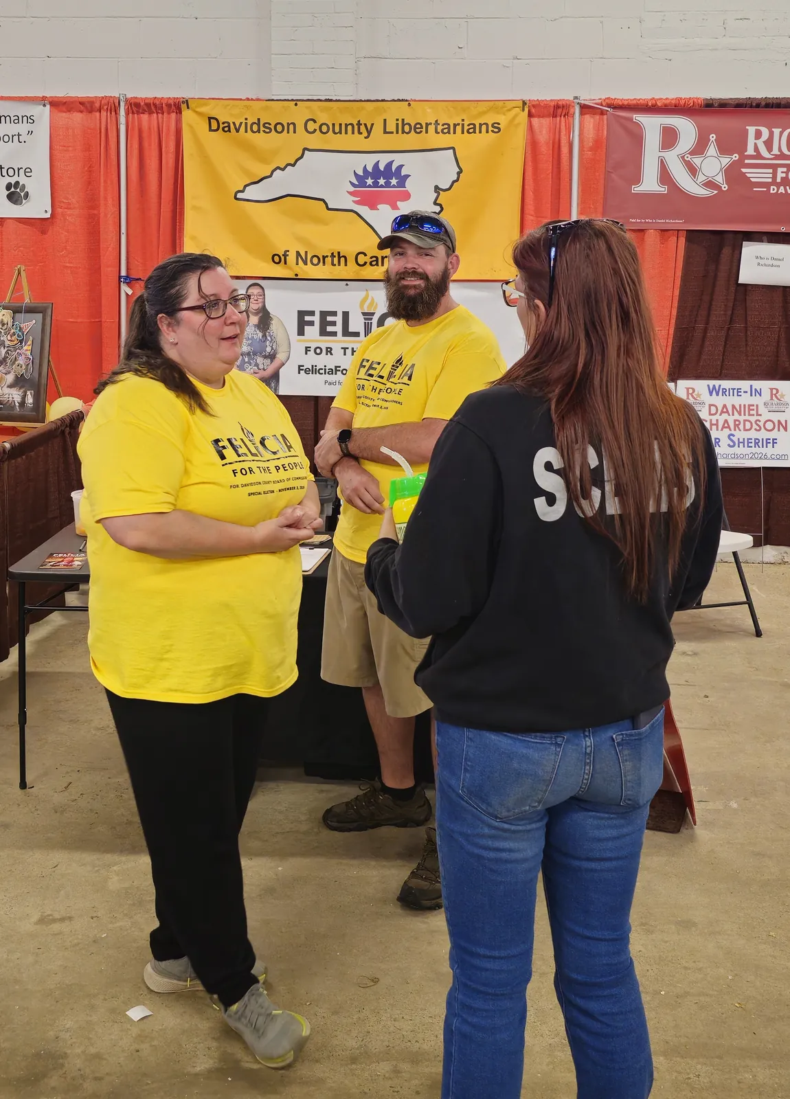 Felicia Dreher in a yellow campaign shirt talking with a volunteer and a visitor at the Davidson County Libertarians booth
