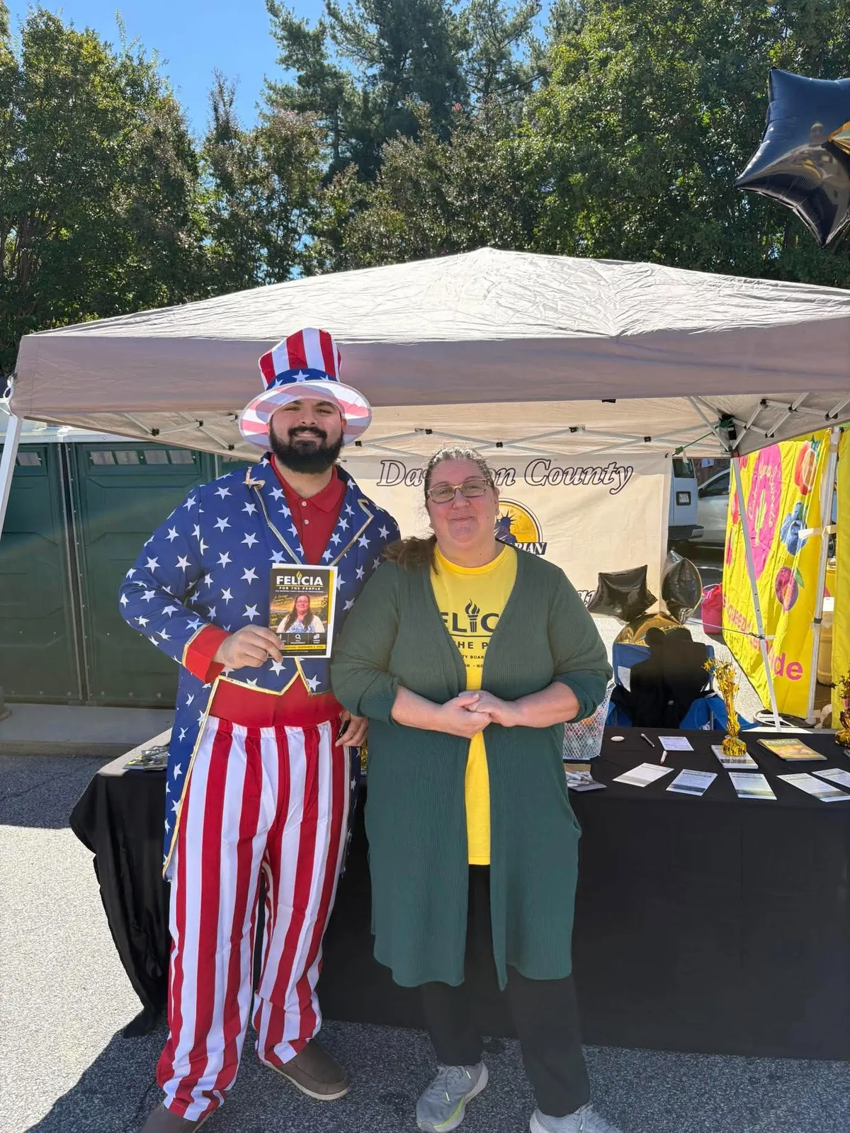 Felicia Dreher standing next to a supporter in a patriotic suit who holds her campaign flyer, in front of the Davidson County Libertarians tent